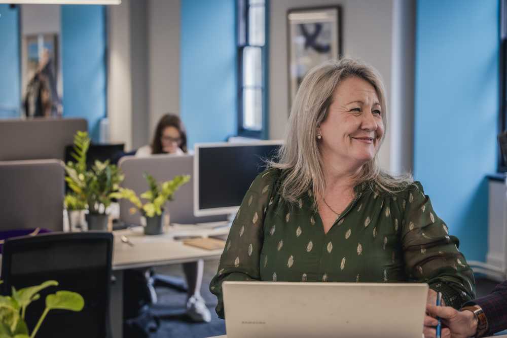 Kathleen smiling, wearing in a green blouse and working at at a laptop