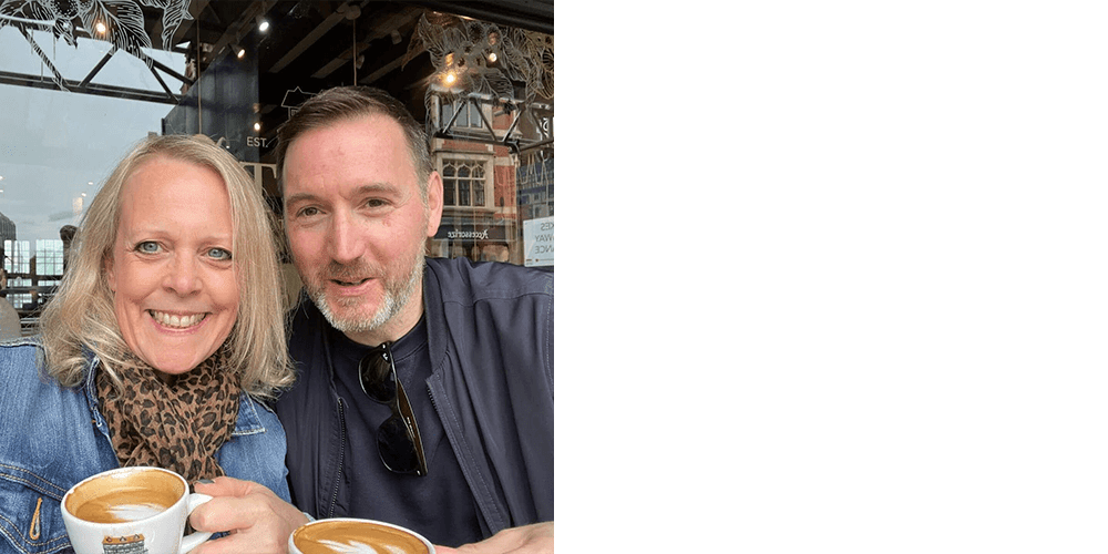 Nicola and Tony smiling at the camera in front of a cafe with coffees in hand.